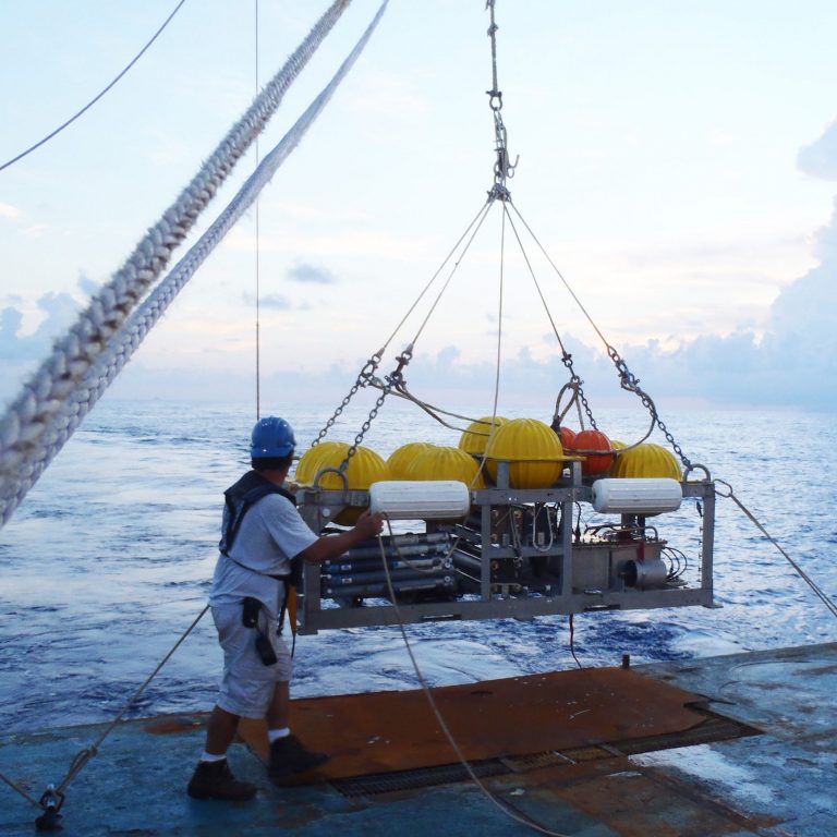 The injection sled is equipped and ready for deployment from the RV Brooks McCall. The white bumpers provide buoyancy for recovery. The grey horizontal tubes, next to engineer Brian Guest, sample salinity to calibrate the CTD behind them. The tracer reservoirs (center) have batteries that run the injectors. The yellow “hard hats” encase hollow glass spheres, making the sled neutrally buoyant as it streams behind a cable when towed at 1 knot. (Photo credit: Laura Spencer)