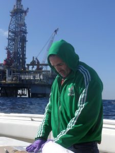 The research team's second sampling expedition in December 2012, was more challenging due to the close proximity of a BP survey vessel, Olympic Triton, which was operating two ROVs, and a mobile offshore deep drilling unit. The rig and vessel each require a reasonable stand-off distance for safety precautions. WHOI's Chris Reddy is shown here, with the drilling rig behind him. (Photo by Christoph Aeppli, Woods Hole Oceanographic Institution)