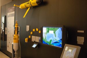 Some of GISR-related research on display at the Bush Library: standing is 1/4 scale replica of an automated monitoring buoy; mounted on the wall is a full-sized ocean glider, GISR Driftcards and bookmarks with an interactive map of the Gulf of Mexico to view their dispersion; and an interactive “Make Your Own Oil Spill” visualization. (Photo courtesy of Bush Library)
