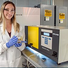 Sydney Niles holds a jacketed beaker containing Macondo oil on water after photo-irradiation in the Atlas solar simulator (right). (Photo credit: Stephen Bilenky)
