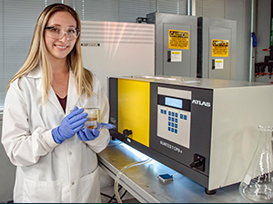 Sydney Niles holds a jacketed beaker containing Macondo oil on water after photo-irradiation in the Atlas solar simulator (right). (Photo credit: Stephen Bilenky)