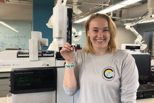 Madison Schwaab, a University of South Florida master’s student, stands in front of the gas chromatography-tandem mass spectrometer holding an almaco jack liver extract. (Provided by Madison Schwaab)